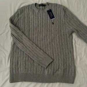 Grå kabelstickad tröja Polo Ralph Lauren - Denna grå kabelstickade tröja från Polo Ralph Lauren kombinerar tidlös stil med hög kvalitet, och erbjuder en mjuk och behaglig känsla tack vare det exklusiva materialet och den sofistikerade kabelstickningen, medan den ikoniska logotypen ger en distinkt touch och den neutrala färgen gör den lätt att matcha med olika outfits, vilket gör den till ett mångsidigt och oumbärligt plagg för alla tillfällen.