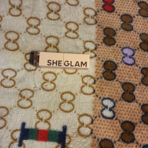 Sheglam läppglans i nude-brun - Snyggt läppglans från Sheglam i en nude-brun nyans med guldig shimmer. Kommer i en stilren fyrkantig förpackning med genomskinligt lock. Perfekt för dig som vill ha glansiga läppar med en hint av färg och skimmer.