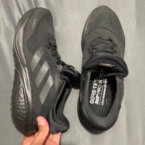 Svarta Adidas sneakers med Gore-Tex - Svarta Adidas sneakers med klassiska tre ränder på sidan och vattentätt Gore-Tex-material. Skorna har snörning, platt sula och är tillverkade i syntet och textil. Perfekta för dig som vill ha stilrena och funktionella skor till vardags eller träning.