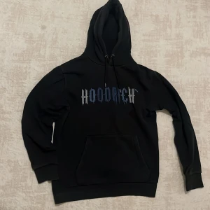 Svart hoodie från Hoodrich med tryck - Svart hoodie från Hoodrich i storlek M med blått och vitt tryck på bröstet. Klassisk känguruficka framtill, snörning i huvan och ribbade muddar. Perfekt för dig som gillar streetwear och vill ha en stilren look.