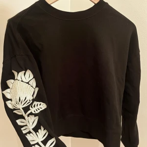 Svart sweatshirt Scotch & Soda XS - Svart sweatshirt från Scotch & Soda i storlek XS. Tröjan har en stor vit broderad blomma längs ena ärmen som ger en unik och cool look. Klassisk rund halsringning och lång ärm. Perfekt för dig som vill sticka ut lite extra.