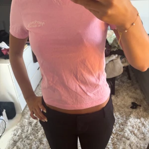 Rosa croppad t-shirt Juicy Couture - Säljer en ljusrosa croppad t-shirt från Juicy Couture med diskret broderad logga på bröstet. T-shirten har rund halsringning och korta ärmar, perfekt för en chill och trendig look.