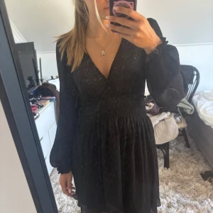 Svart kort omlottklänning med v-ringning - Supersnygg svart kort klänning med v-ringning och långa, lätt transparenta ärmar. Smockad midja som ger en fin siluett och omlottkänsla upptill. Perfekt för dig som gillar en klassisk men trendig look.