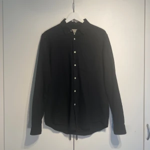 Svart skjorta från L'Homme Rouge - Stilren svart skjorta från L'Homme Rouge med klassisk krage och vita knappar framtill. Skjortan har en bröstficka och är långärmad. Materialet är mjukt och ser ut att vara bomull, perfekt för en clean och enkel look. Storlek M och true to size. Avslappnad passform, perfekt för sommaren. 