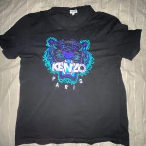 Svart Kenzo t-shirt med tigertryck - Svart t-shirt från Kenzo med ikoniskt broderat tigerhuvud i blått och lila på bröstet samt Kenzo Paris-tryck. Klassisk passform och rund hals. Tillverkad i mjuk bomull, perfekt för dig som vill sticka ut med en fet logga. Dock har en sak inuti tröjan låsnat lite.