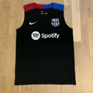 FC Barcelona svart ärmlös fotbollströja Nike - Säljer en svart ärmlös FC Barcelona fotbollströja från Nike med Dri-FIT-material. Tröjan har klubbmärke och Nike-logga på bröstet, samt Spotify som sponsor. Kragen har röda och blå detaljer. Perfekt för träning eller match. Och så har shortsen djupa fickor 
