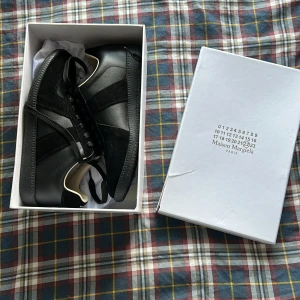 Maison Margiela Replica svarta sneakers - Maison Margiela Replica sneakers i helsvart med både skinn och mocka-paneler. Klassisk låg modell med rund tå, svarta snören och diskret branding på insidan. Snygg minimalistisk look som passar till allt. Kommer med originalkartong och dustbags. Lite skada på lådan.