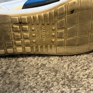 Valentino Open sneakers vit/blå skinn - Snygga Valentino Open sneakers i vitt skinn med bred blå skinnpanel över mitten. Klassisk låg modell med rund tå, vita snören och diskret logga på plösen. Perfekta för dig som gillar exklusiva och stilrena sneakers med en twist.