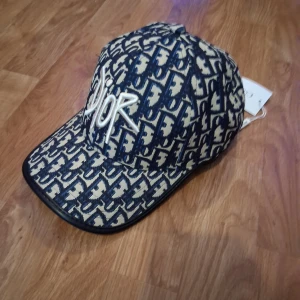 Dior keps med monogrammönster - Exklusiv keps från Dior med ikoniskt blått och beige monogrammönster och broderad Dior-logga framtill. Justerbar rem baktill med metallspänne. Snygg och trendig accessoar som lyfter vilken outfit som helst.