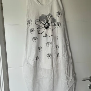 Vit långklänning med blommigt tryck - Luftig vit långklänning med stora och små svarta blommor framtill. Ärmlös modell med rund halsringning och flowy passform. Perfekt för varma dagar och har en avslappnad, bohemisk vibe.