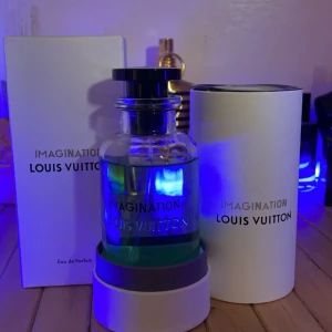Louis Vuitton Imagination Parfym - En elegant parfymflaska från Louis Vuitton med en transparent, ljusgrön vätska. Flaskan är rundad med en svart topp och har tydlig Louis Vuitton-logga på framsidan. Den kommer i en vit kartong med svart text. Perfekt för den som gillar lyxiga och stilrena dofter. Flaskan är av glas och har en modern, minimalistisk design som utstrålar exklusivitet.