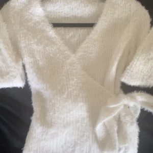 Vit fluffig omlottkofta med knytband - Supermjuk och fluffig vit omlottkofta med knytband i midjan. Koftan har lång ärm och är gjord i ett stickat material som känns riktigt mysig . Väldigt mjuk i materialet . Och strechig så passar många storlekar . Aldrig använd .