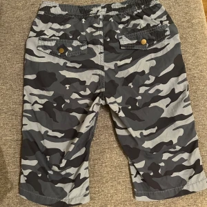 Camo shorts med snörning - Snygga camouflagemönstrade shorts i svart, grått och vitt med elastisk midja och snörning framtill. Shortsen har sidofickor och bakfickor med knappdetaljer. Perfekta för en avslappnad streetstil under sommaren.