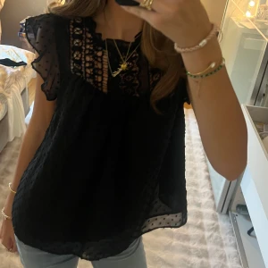 Svart blus med spets och volang - Supersöt svart blus med transparenta prickiga ärmar och volangdetaljer. Ovanför bysten finns ett parti i spets. Blusen är luftig och har korta ärmar, perfekt med jeans eller kjol.