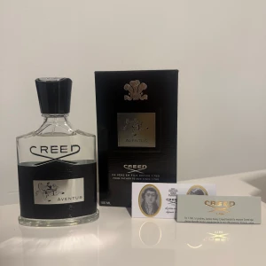 Creed Aventus 100ml Eau de Parfum - Lyxig parfym från Creed, Aventus 100ml. Flaskan är genomskinlig med svart och silverdetaljer, och har en svart kork. Kommer med original svart kartong och äkthetskort. Doften är ikonisk och exklusiv, perfekt för dig som gillar eleganta och fräscha parfymer.