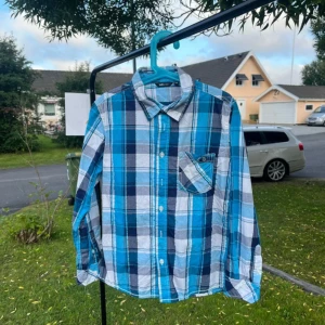 Blå rutig skjorta från Lindex Kids - Snygg blå och vit rutig skjorta från Lindex Kids i storlek 140. Skjortan har klassisk krage, lång ärm och en bröstficka med knapp. Perfekt för dig som gillar en avslappnad men ändå stilren look.