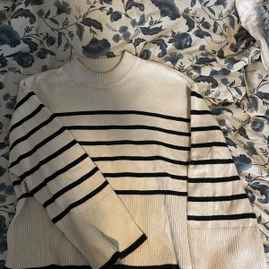 Randig stickad tröja från Gina Tricot - Superfin stickad tröja från Gina Tricot i vitt med svarta ränder. Tröjan har breda ribbade muddar, hög hals och lång ärm. Perfekt för lager-på-lager och ger en clean, trendig look.