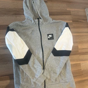 Grå zip-hoodie från Nike Air Tech - Grå hoodie från Nike Air i storlek XL med hel dragkedja, huva och fickor. Ärmarna har vita och svarta detaljer som ger en sportig vibe. Perfekt för chill dagar eller när du vill ha en clean streetstyle-look. Storlek XL FÖR BARN eller 158-170