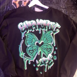 Svart sweatshirt med grön fjärilsgrafik - Cool svart sweatshirt med stor grön och turkos fjärilsgrafik och graffiti-texten 'Euphoric Life' på framsidan. Tröjan har rund hals och långa ärmar, perfekt för dig som gillar streetstyle och unika prints.