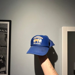 Blå truckerkeps Buckhannon Stockyards - Snygg blå truckerkeps med vit och gul brodyr framtill, där det står Buckhannon Stockyards och har motiv av kor. Kepsen har mesh på sidorna och justerbar baksida. 