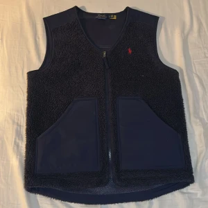 Mörkblå fleeceväst Polo Ralph Lauren - Snygg mörkblå fleeceväst från Polo Ralph Lauren med röd broderad logga på bröstet. Västen har två stora fickor framtill och dragkedja. Perfekt lager-på-lager-plagg med mjuk teddyfleece och sportig känsla.