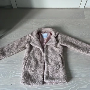 Beige teddyjacka från 157, stl 130/140 - Supermjuk och fluffig teddyjacka i beige från 157. Jackan har långa ärmar, två fickor framtill och en klassisk krage. Perfekt för dig som vill ha en mysig och trendig look under kyliga dagar. Fodrad insida i ljusrosa tyg för extra komfort.