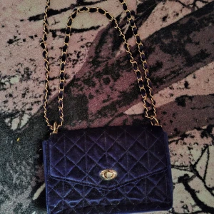 Mörkblå quiltad axelväska Holly & Whyte - Säljer en mörkblå quiltad axelväska från Holly & Whyte i sammet. Väskan har guldfärgad kedjerem och snyggt vridlås framtill. Insidan har ett fack med dragkedja och en metallbricka med logga. Perfekt för dig som vill ha en stilren och trendig väska.