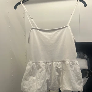 Vit volangig linnetopp - Supersöt vit linnetopp med tunna axelband och volangig nederdel. Perfekt för varma dagar när du vill ha en luftig och enkel stil. Toppens material känns mjukt och lätt mot huden.