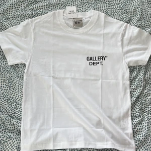 Vit t-shirt från Gallery Dept. - Vit t-shirt från Gallery Dept. med svart tryck fram och stort tryck på ryggen. Klassisk passform, rund hals och korta ärmar. Tillverkad i mjuk bomull, perfekt för en clean och stilren look.
