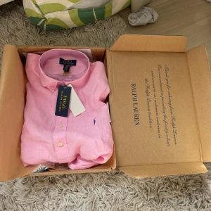 Rosa linneskjorta från Ralph Lauren (NY) - Snygg rosa skjorta från Ralph Lauren i mjukt linne. Klassisk button-down krage, broderad logga i blått på bröstet och knäppning framtill. Perfekt för dig som vill ha en fräsch och stilren look med en touch av färg.