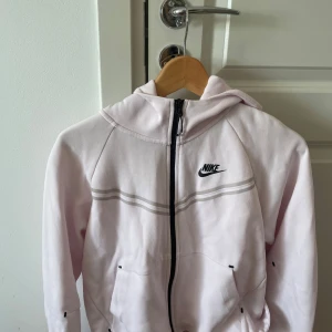 Ljusrosa Nike hoodie med dragkedja - Säljer en ljusrosa hoodie från Nike med hel dragkedja och huva. Tröjan har svarta Nike-loggan på bröstet, två fickor framtill och en snygg randig detalj över bröstet. Perfekt för chill dagar eller när du vill ha en sportig look.