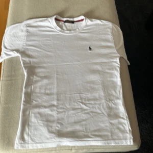 Vit t-shirt från Polo Ralph Lauren - Klassisk vit t-shirt från Polo Ralph Lauren med rund hals och broderad marinblå logga på bröstet. Tillverkad i mjuk bomull som känns skön mot huden. Perfekt basic-plagg med snygg passform och stilren look.