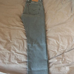 Grå Levis 501 straight jeans - Säljer ett par klassiska grå Levis 501 jeans med raka ben och normal passform. Jeansen har fem fickor, knappgylf och den ikoniska Levis-lappen bak. Perfekta för dig som gillar en tidlös look och vill ha ett par jeans som funkar till allt.