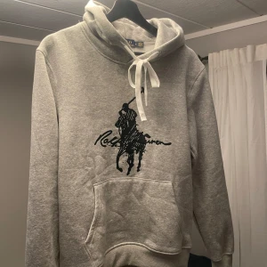 Grå hoodie från Polo Ralph Lauren - Snygg grå hoodie från Polo Ralph Lauren med stor svart logga framtill. Klassisk känguruficka, vit snörning i huvan och mjukt material som känns skönt mot huden. Perfekt för dig som gillar stilren streetwear med premiumkänsla.