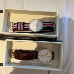 Daniel Wellington klockor med olika armband - Två stilrena Daniel Wellington klockor med vit urtavla. En har ett randigt tygarmband i blått, rött och vitt, medan den andra har ett klassiskt brunt läderarmband och guldfärgad boett. Batteri saknas, men box tillkommer. 150 kr styck, 300 kr för båda. 