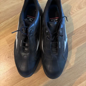 Svarta golfskor från Mizuno - Snygga svarta golfskor från Mizuno med vita detaljer och klassisk snörning. Skorna har en robust sula med röda dobbar för grymt grepp. Tillverkade i skinn för bra känsla och passform.