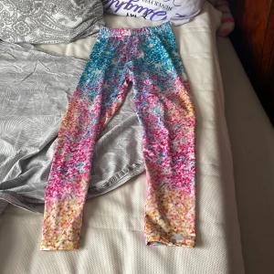 Färgglada leggings med mönster - Säljer ett par leggings i storlek 140 med färgstarkt mönster i blått, rosa, gult och vitt. De är tillverkade i stretchigt material (95% polyester, 5% spandex) och har resår i midjan för skön passform. Perfekta för dig som gillar att sticka ut med din stil!