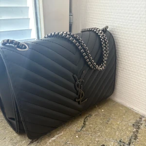 Svart quiltad axelväska från YSL -  Snygg svart axelväska från YSL med quiltad design och kedjerem i metall. Väskan har YSL-logga framtill och ett innerfack med dragkedja. Perfekt storlek för det viktigaste och ger en lyxig känsla till din outfit.