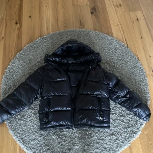 Svart pufferjacka Polo Ralph Lauren - Snygg svart pufferjacka från Polo Ralph Lauren med glansig finish och röd broderad logga på bröstet. Jackan har huva, dragkedja framtill och två fickor med dragkedja. Perfekt för kalla dagar och riktigt stilren.Jackan kostade 4000 och är knappast använd då den inte passade.