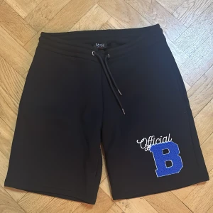 Svarta shorts BoohooMAN med blå patch - Svarta shorts från BoohooMAN med snörning i midjan och en stor blå patch med texten 'Official B' på ena benet. Sköna och avslappnade, perfekta för chill dagar. Materialet känns mjukt och sportigt.