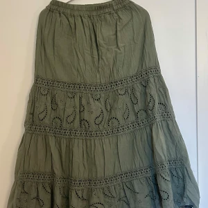 Olivgrön långkjol med broderade detaljer - Olivgrön långkjol i bomull med broderade paisleymönster och virkade spetsband. Kjolen har resår i midjan och ett flowy fall, perfekt för en chill och bohemisk vibe. Snygga detaljer med hålbroderi och spets ger kjolen en unik look.