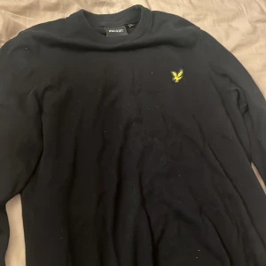 Svart stickad tröja från Lyle & Scott - Svart stickad tröja från Lyle & Scott med klassisk gul logga på bröstet. Tröjan har rund halsringning och långa ärmar, perfekt för lager-på-lager-stil. Enkel och stilren design som funkar till det mesta. Knappt använd. 