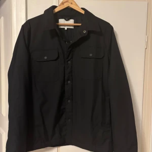 Svart overshirt med knappar - Stilren svart overshirt med klassisk krage och tryckknappar framtill. Två bröstfickor med lock och knäppning ger en clean look. Perfekt att slänga över en hoodie eller t-shirt för en chill vibe.