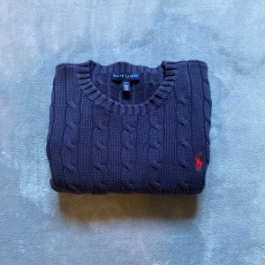 Mörkblå stickad tröja från Ralph Lauren - Snygg mörkblå kabelstickad tröja från Ralph Lauren med klassisk rund halsringning och röd broderad logga på bröstet. Tröjan har långa ärmar och ribbade muddar för en clean och tidlös look. Perfekt för lager-på-lager under kyliga dagar. Storlek S men passar XS. Tveka inte att höra av dig vid frågor. 