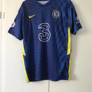 Chelsea FC matchtröja Nike blå 176 - Chelsea FCs officiella matchtröja från Nike i blått med gula detaljer. Tröjan har korta ärmar, Dri-FIT-material och ett unikt mönster med både zigzag och rutor. Klubbmärke, sponsorloggor och Nike Swoosh på bröstet. Perfekt för fotbollsfans!