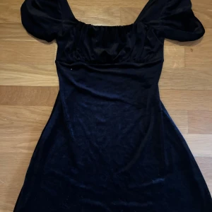 Svart sammetsklänning med puffärm - Supersnygg svart sammetsklänning med kort längd och puffiga kortärmar. Klänningen har en söt, rynkad byst och en lätt utsvängd kjol. Perfekt för dig som gillar en klassisk men trendig look.