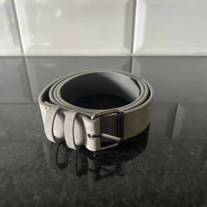 Mockabälte grått  - Snyggt och stilrent grått bälte i mocka med klassiskt metallspänne. Perfekt accessoar för att lyfta din outfit med en clean look. Bältet har en enkel design utan mönster och passar till många olika stilar.