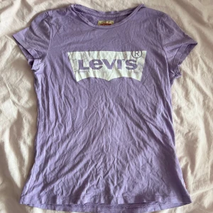Lila Levi's t-shirt med logga - Snygg lila t-shirt från Levi's med klassisk vit logga framtill. T-shirten har korta ärmar och rund halsringning. Tillverkad i mjuk bomull som är skön mot huden. Storleken är i barn men skulle säga att den passar S. 