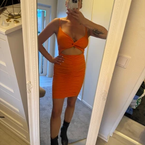 Orange tight kortklänning med cutout - Snygg orange kortklänning med smala axelband, knutdetalj framtill och cutout under bysten. Klänningen är tight, har rynkade sömmar både fram och bak och är tillverkad i ett stretchigt material. 
