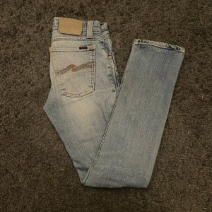 Nudie jeans  - Snygga ljusblå jeans från nudie med klassisk femficksdesign och subtila slitningar. De har en normal passform och är perfekta för en avslappnad stil. Jeansen har en diskret logga på bakfickan och en bekväm midja. W27 l30, super snygga och super sköna. Har två lagade hål som är snygga slitningar 🤩🤩🤩